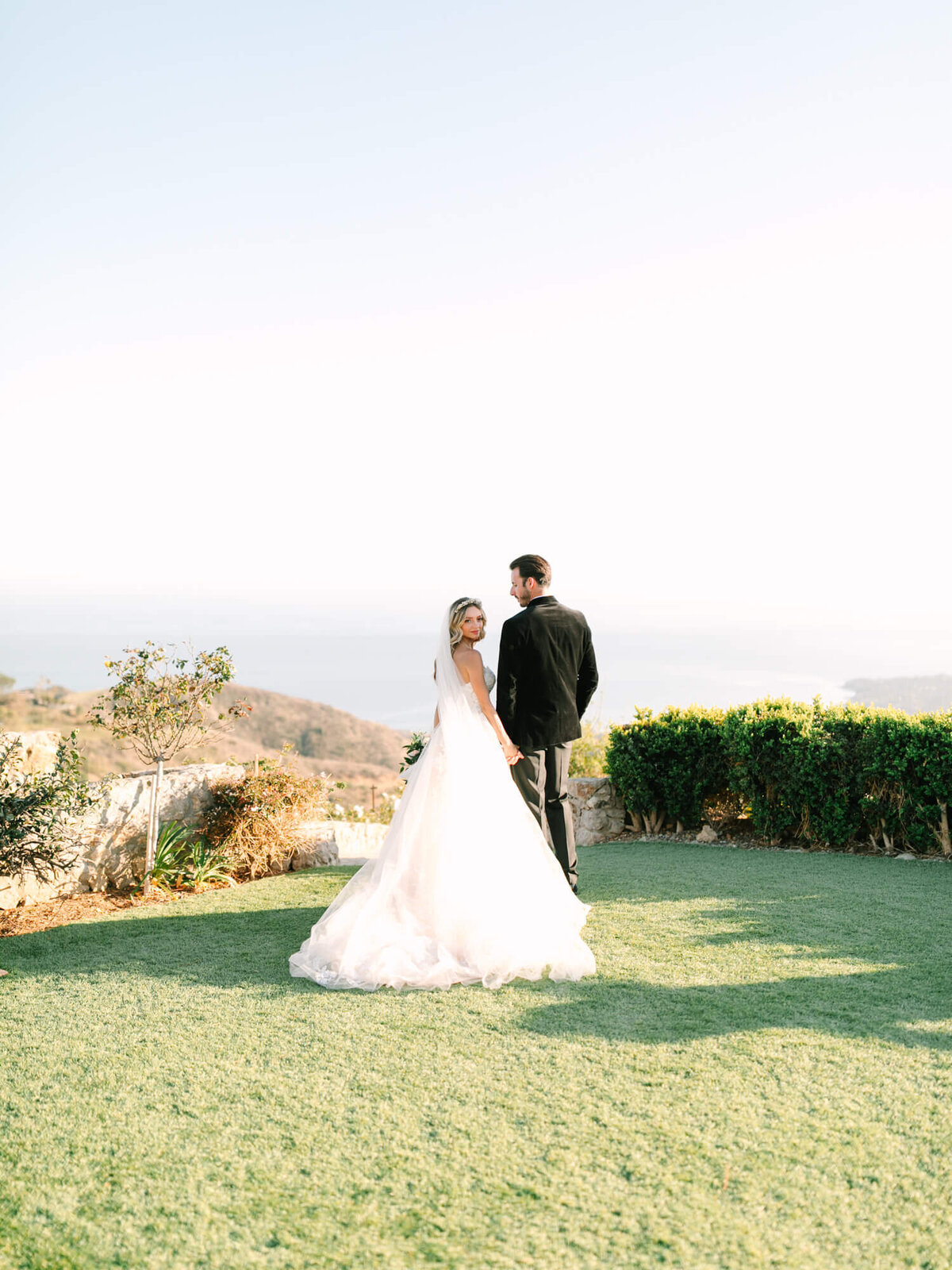 A bride and groom stand on a lush green lawn, overlooking a scenic, sunlit landscape.