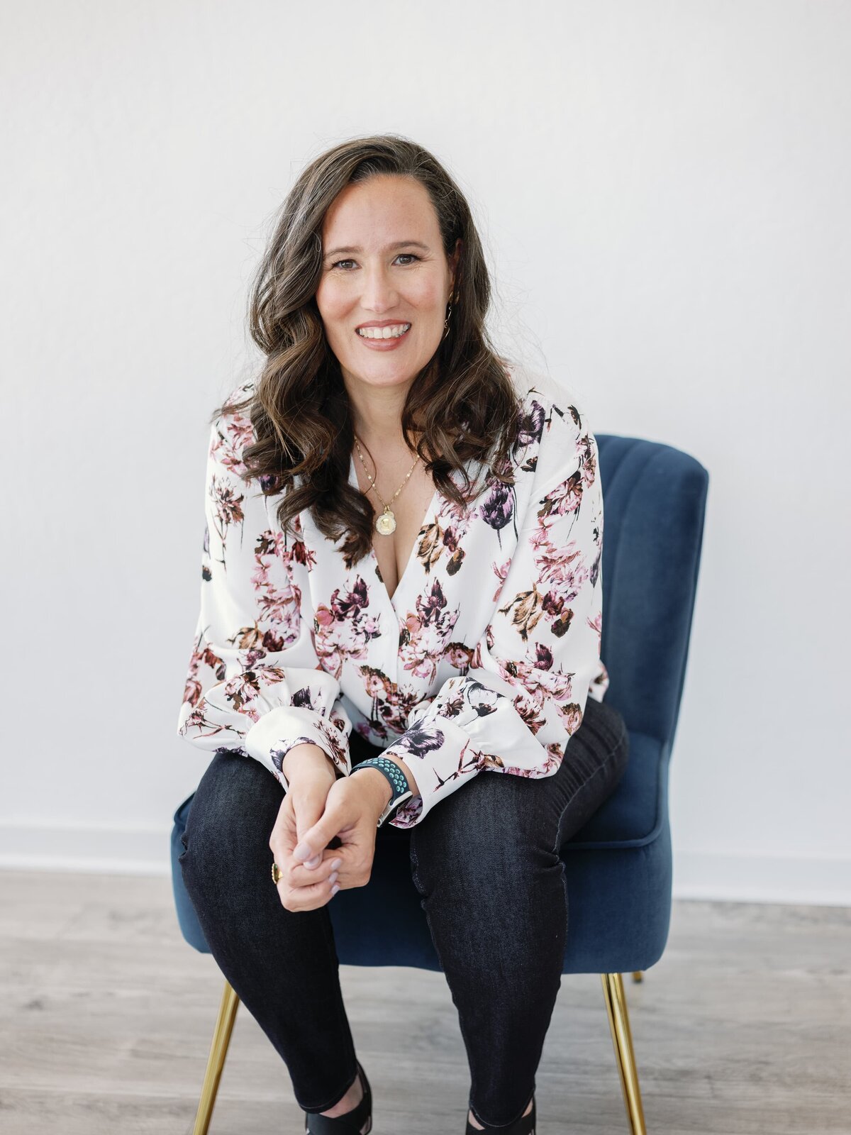 Woman sitting in a blue velvet chair smiling with her hands clapsed and looking at the camera