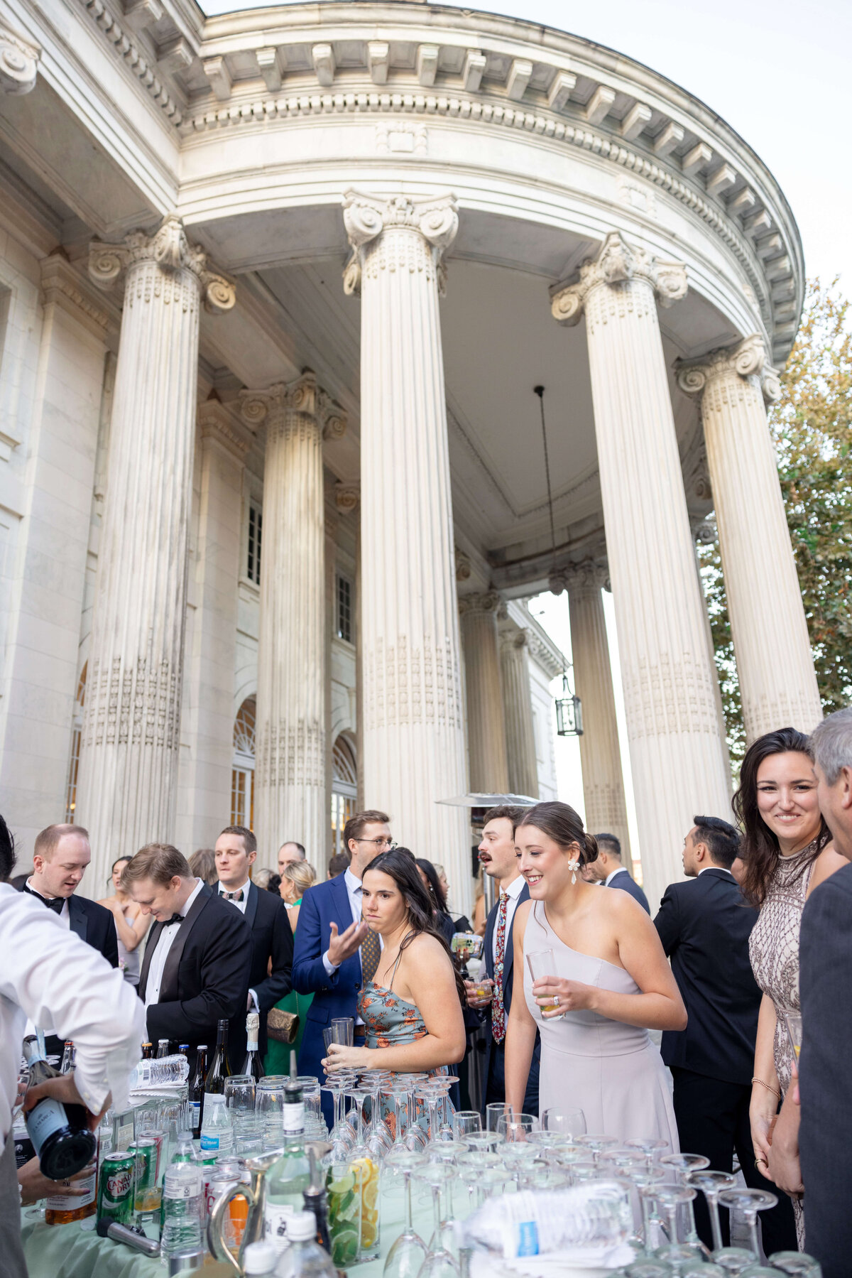 A group of elegantly dressed people gathers at an outdoor bar setup in front of a grand building with tall columns. Tables are filled with drinks and bottles, and the event appears festive and lively.