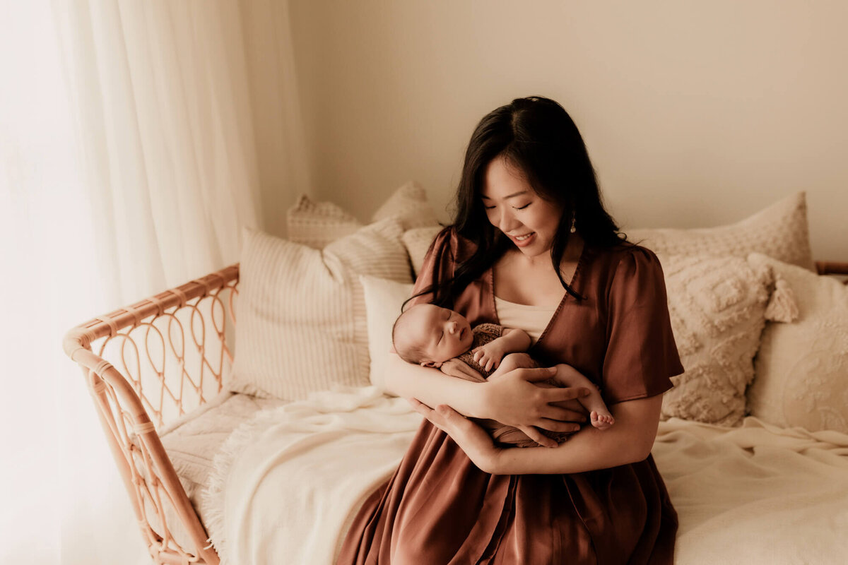 Mother holds her newborn baby boy while sitting on a rattan daybed while wearing a rose colored dress.