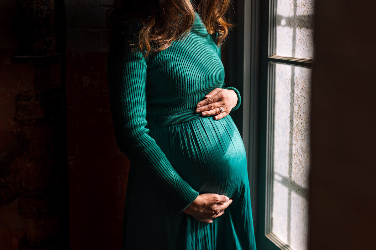 pregnant woman in green dress standing by window holding baby bump at Fort Point in San francisco
