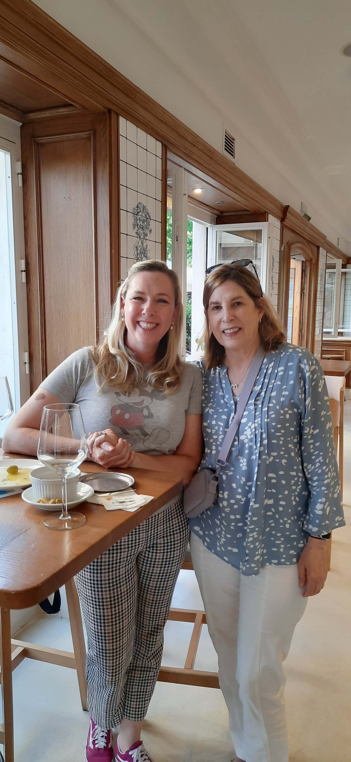 Cristina Barrero and a woman standing next to a table with a wine glass and food while smiling at the camera.