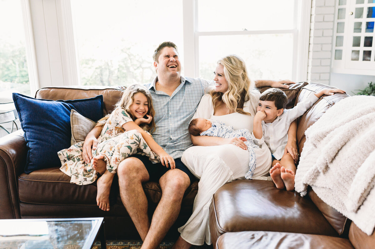 family of five sits on leather couch with newborn at home and dad is laughing