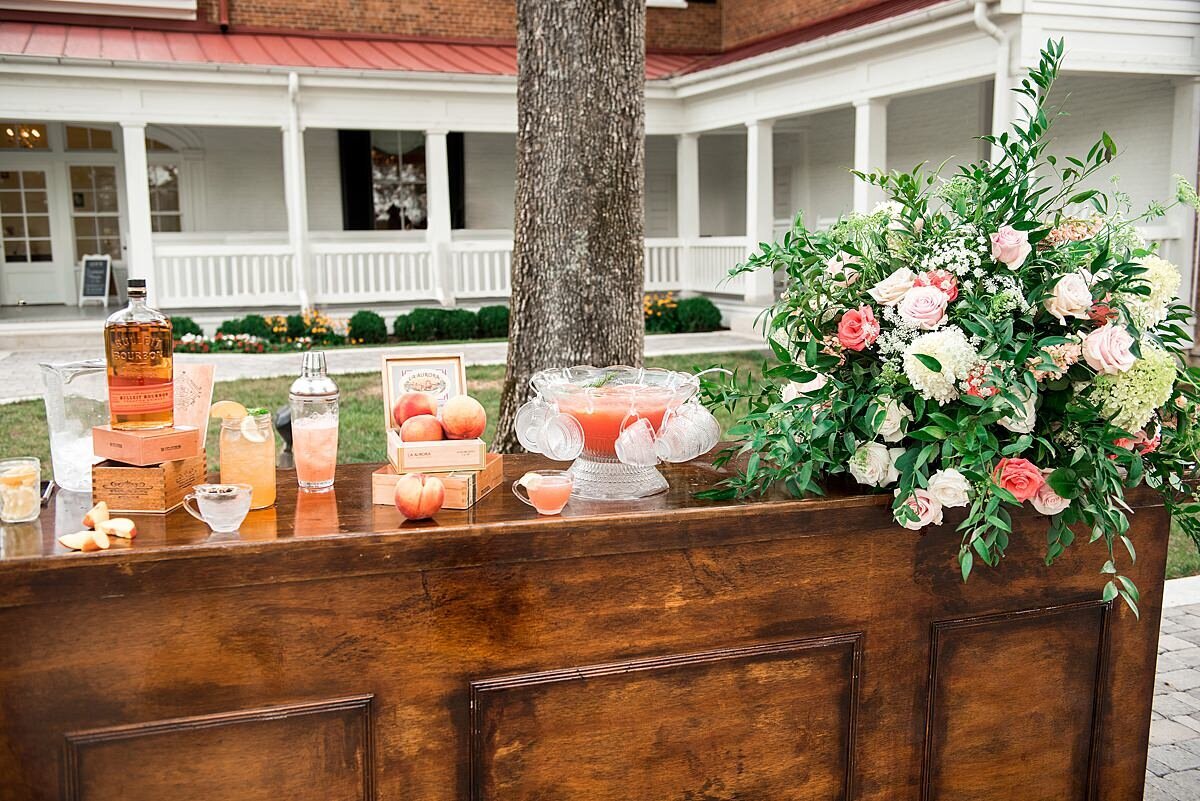 A large dark wood bar decorated with a large centerpiece of greenery, peach and pink, white and ivory flowers.  The bar is topped with an assortment of signature summer cocktails  and signature summer mocktails as well as flavored wedding teas.