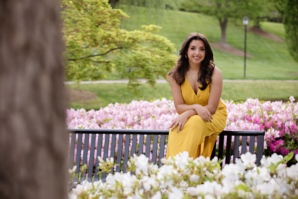 Stunning high school senior in yellow dress seated on a park bench surrounded by white azaleas in front and vibrant pink blooms behind.