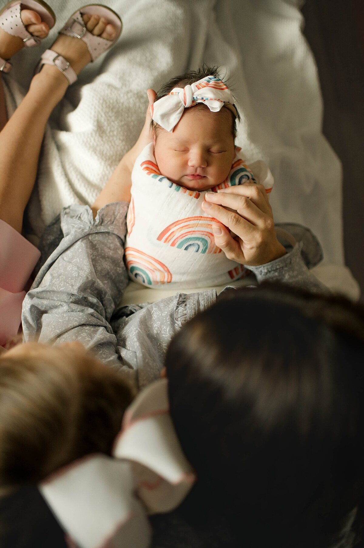 Baby swaddled in rainbow blanket sitting on moms lap with big sister looking at her Houston Newborn Photography session