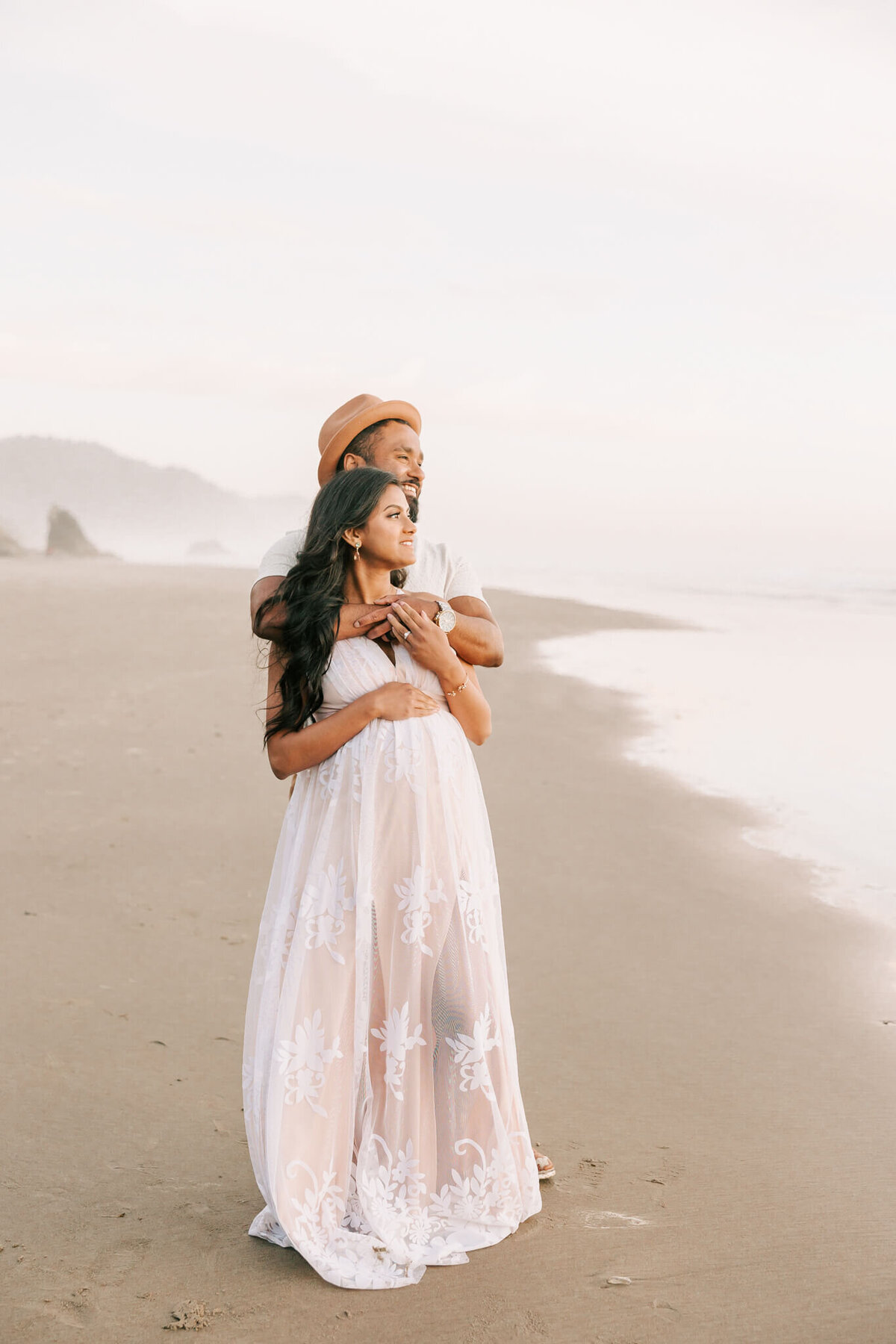 Portrait of a husband hugging his wife. They are looking out at the ocean and she has her hand on her belly and is smiling.