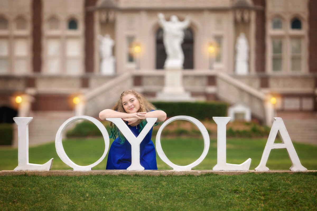 Senior female posing with the LOYOLA sign on the campus of Loyola University in New Orleans.  She is kneeling over the Y in a royal blue dress.  Her head is resting on the hands, which are propped up on the Y.