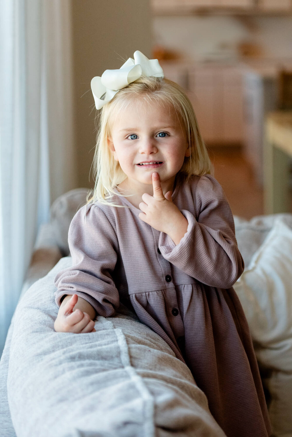 A young toddler girl with a finger on her chin stands on a couch in a window