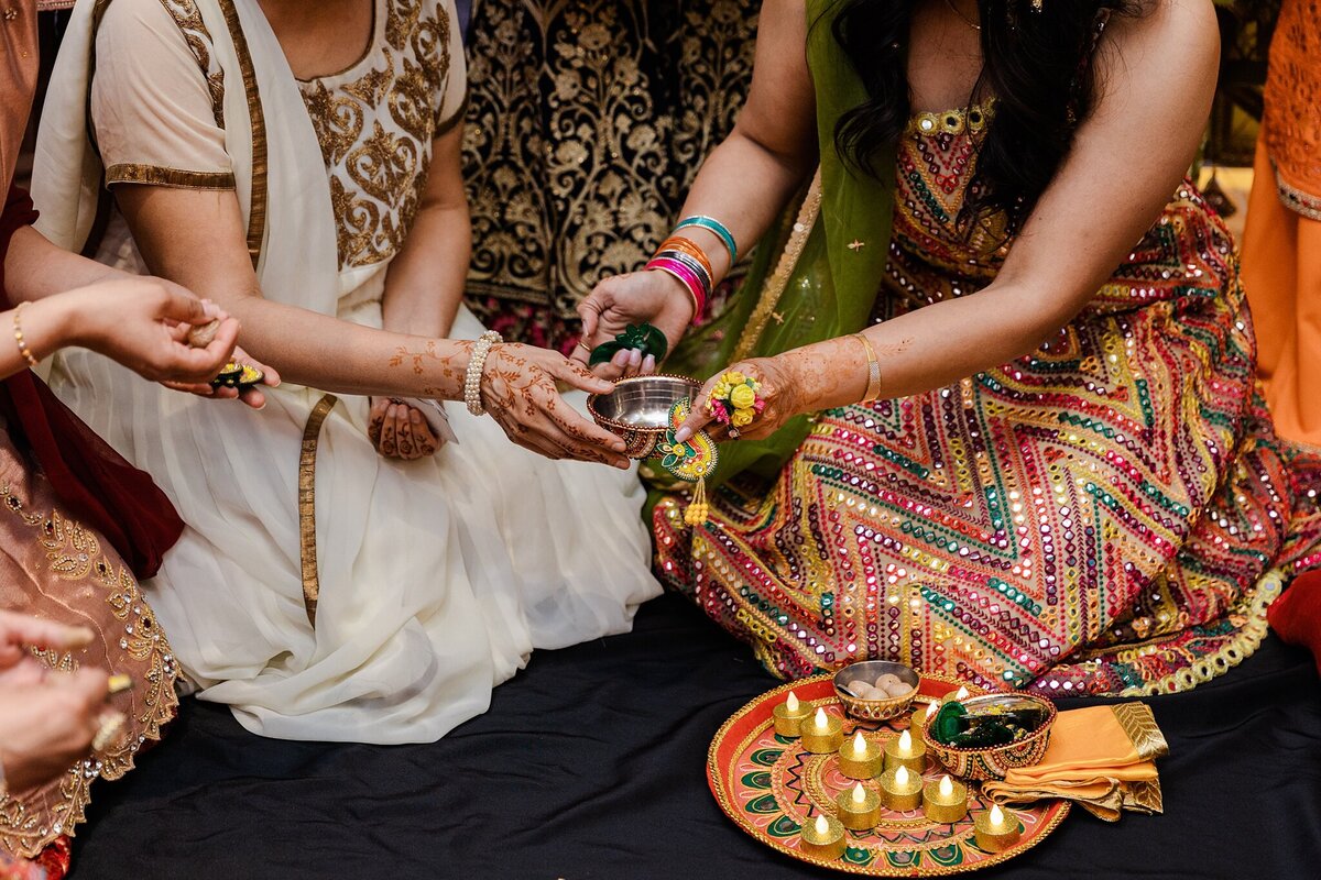 A candid shot of a Pithi ceremony during a wedding in DFW, Texas. Multiple torsos can be seen interacting with the elements of the ceremony while everyone is dressed in traditional wedding attire.
