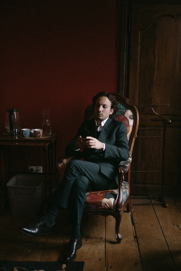 Un garçon d'honneur en costume sombre assis dans un fauteuil ancien, plongé dans son téléphone dans un salon au Château de la Rouerie. Reportage photo Kamescop, spécialiste du mariage en Bretagne.
