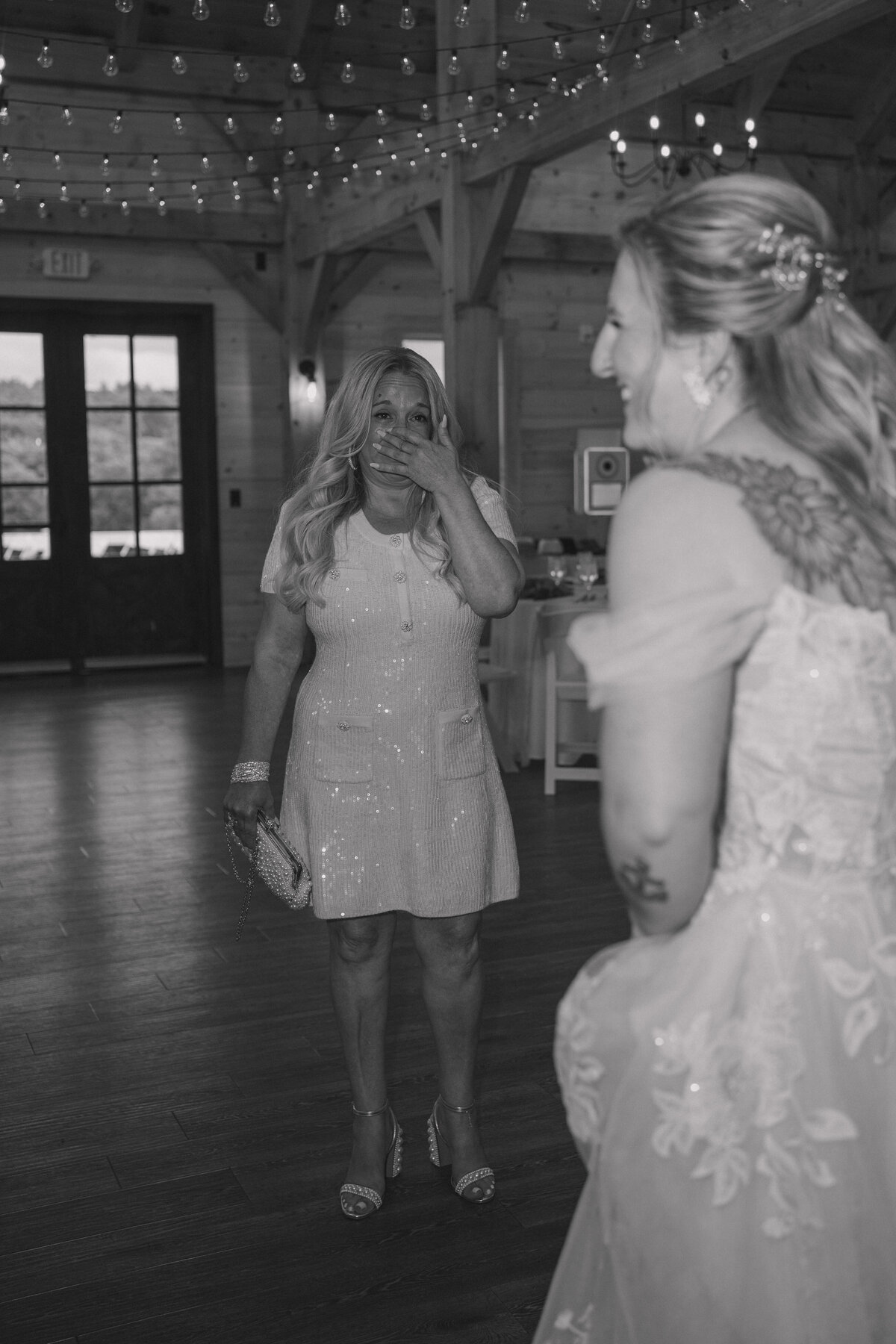 Black and white photo of an emotional first look between a bride and her mother inside a rustic wedding venue adorned with string lights. The mother, wearing a sequined dress, covers her mouth in surprise and joy as she sees her daughter in her wedding gown