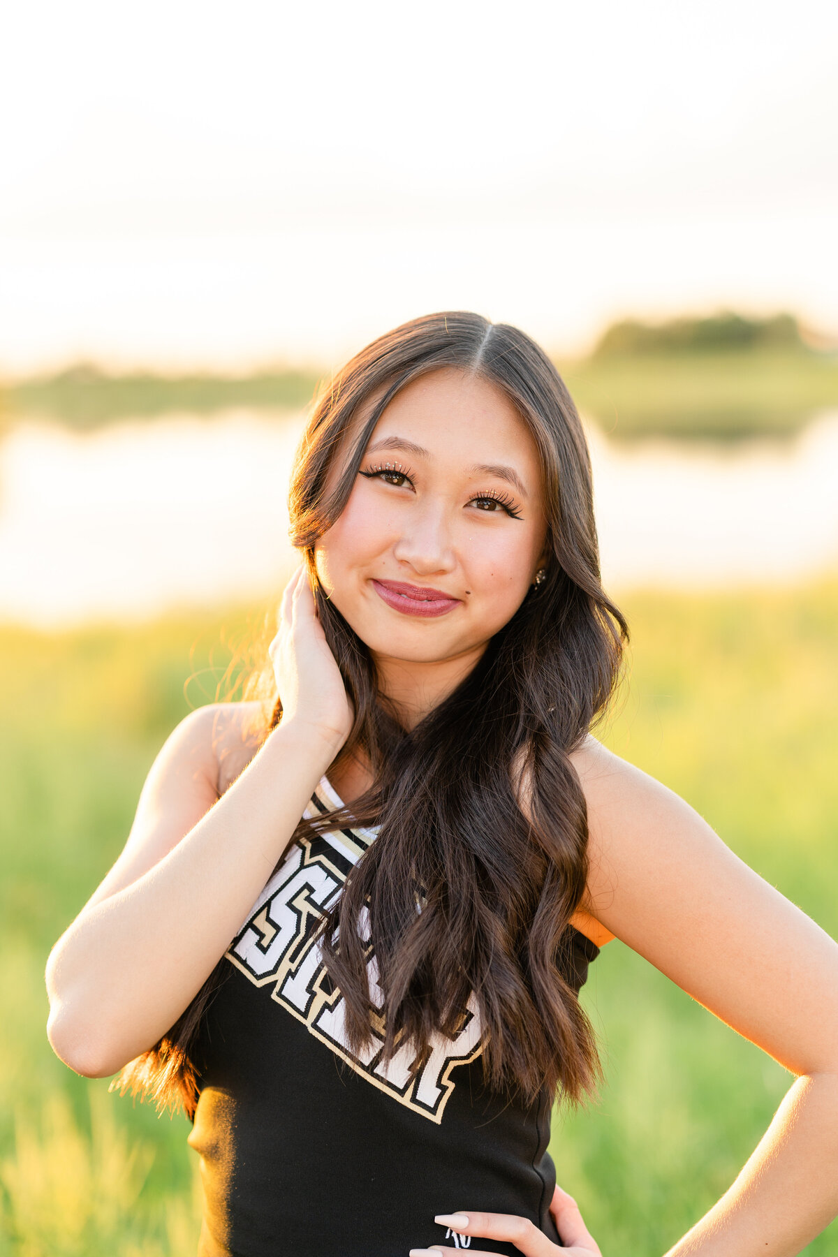Houston high school senior girl in cheerleading outfit holding hair and smiling with hand on hip in open field with pond at golden hour in Josey Lake Park