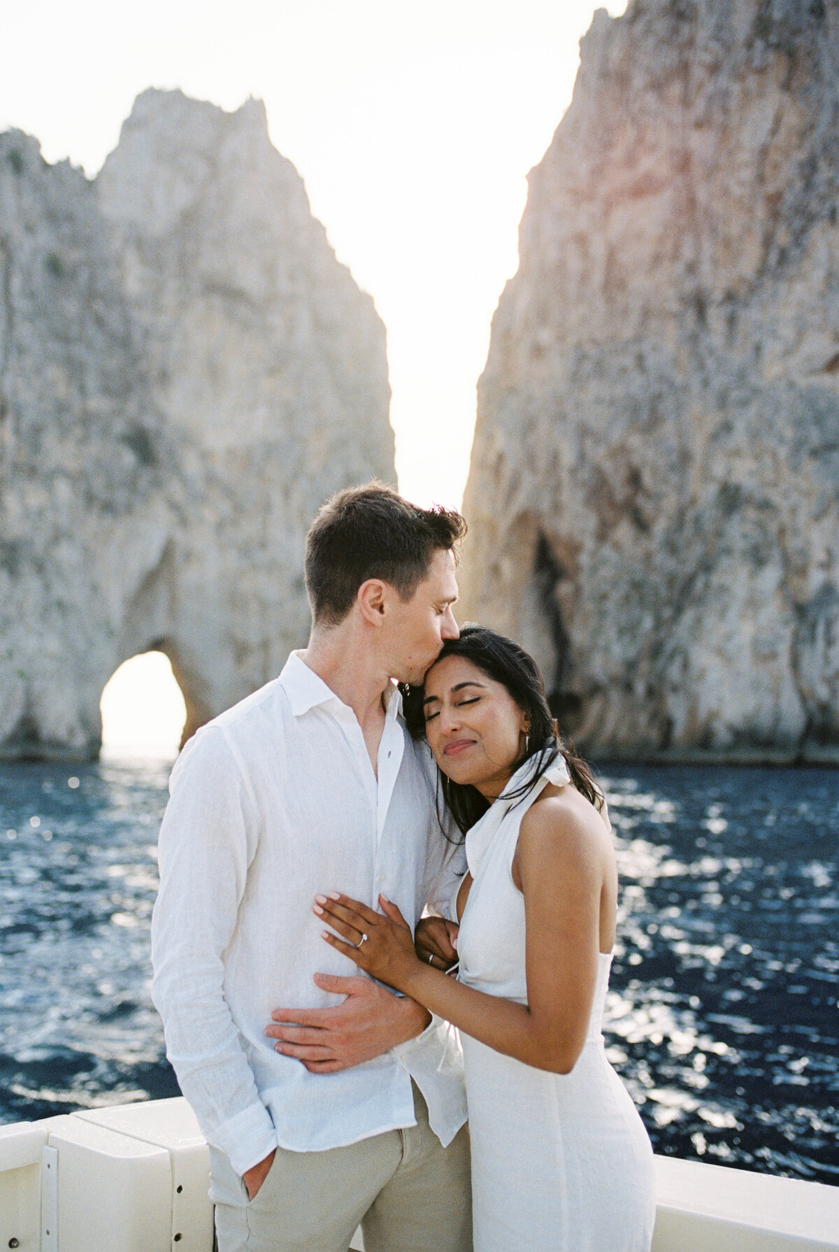 35mm film photo of Romantic couple embracing on a boat with the iconic Faraglioni rock formations of Sorrento in the background, captured during a destination wedding shoot at sunset.