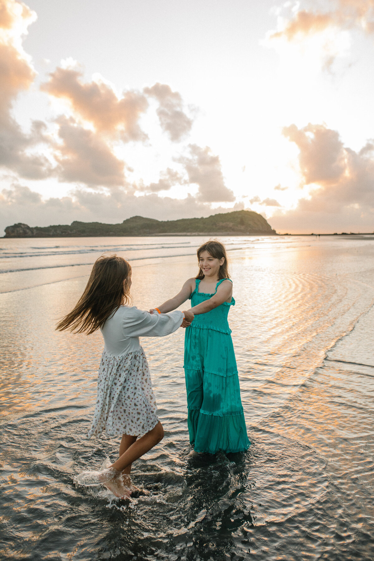 Siblings dance together, holding hands, with shallow waves beneath their feet on the shore.