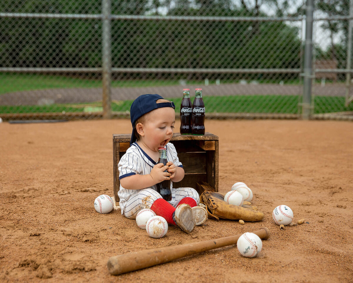 1 year old boy sitting on a baseball diamond with balls, bat and glove around him. He's trying to drink a bottle of coke.