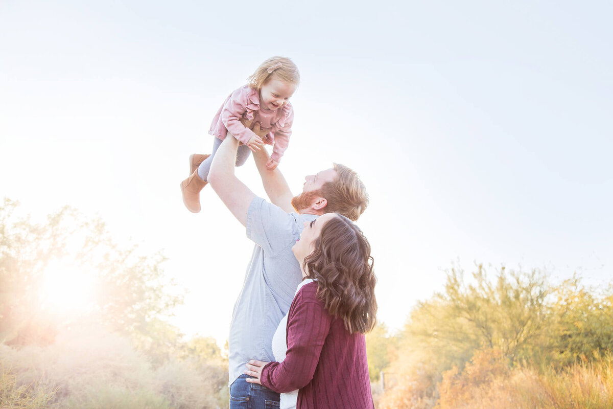 Redheaded man in light blue shirt holding redheaded toddler in pink dress up in the air with wife in maroon looking up laughing