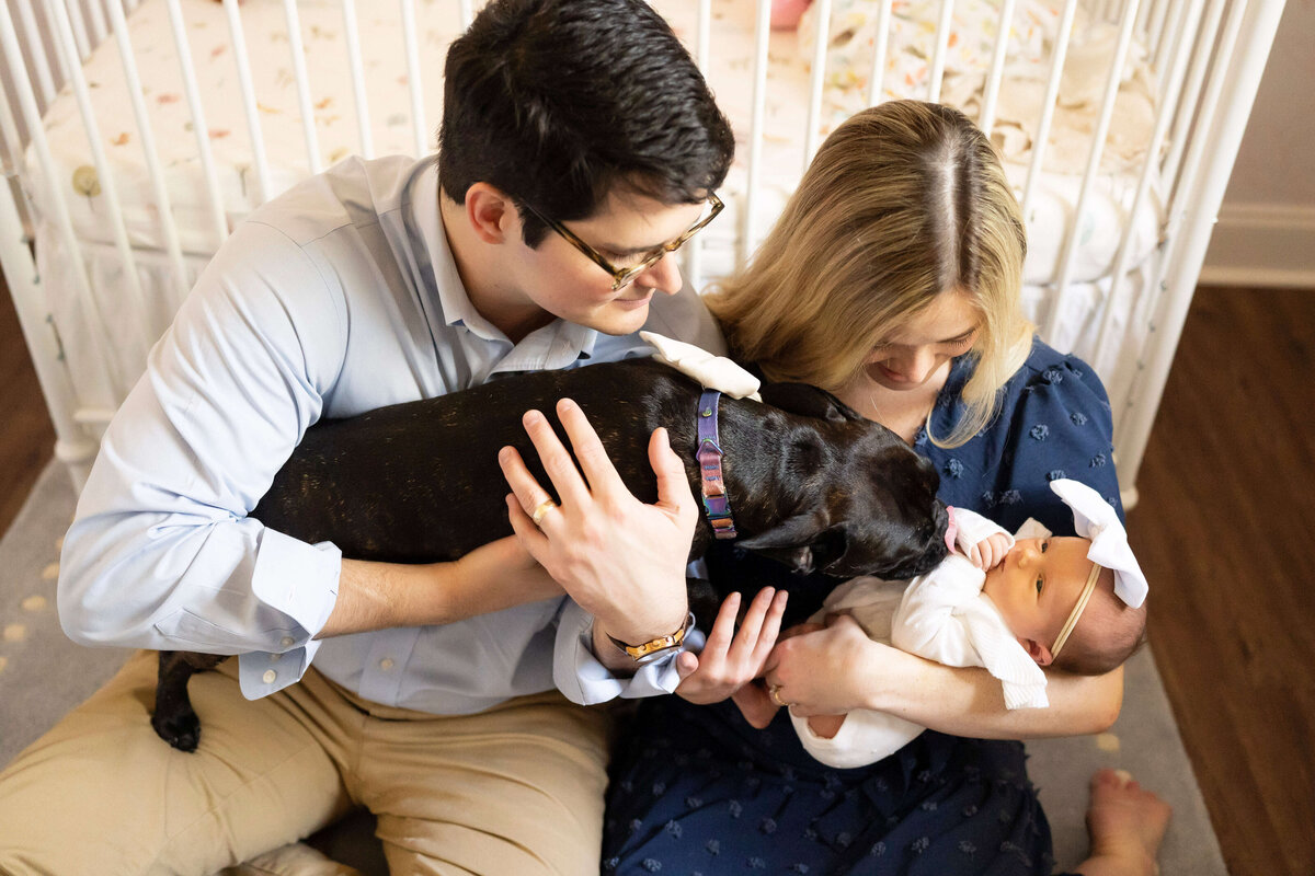 Parents sitting in front of crib holding newborn girl and dog