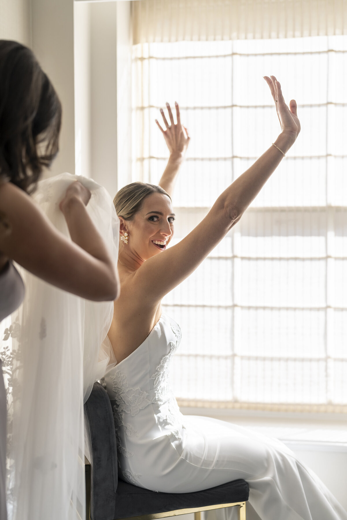 A bride in a white dress sits on a chair with her arms raised joyfully, smiling at the camera. A woman stands beside her adjusting the veil. Sunlight streams through a window in the background.