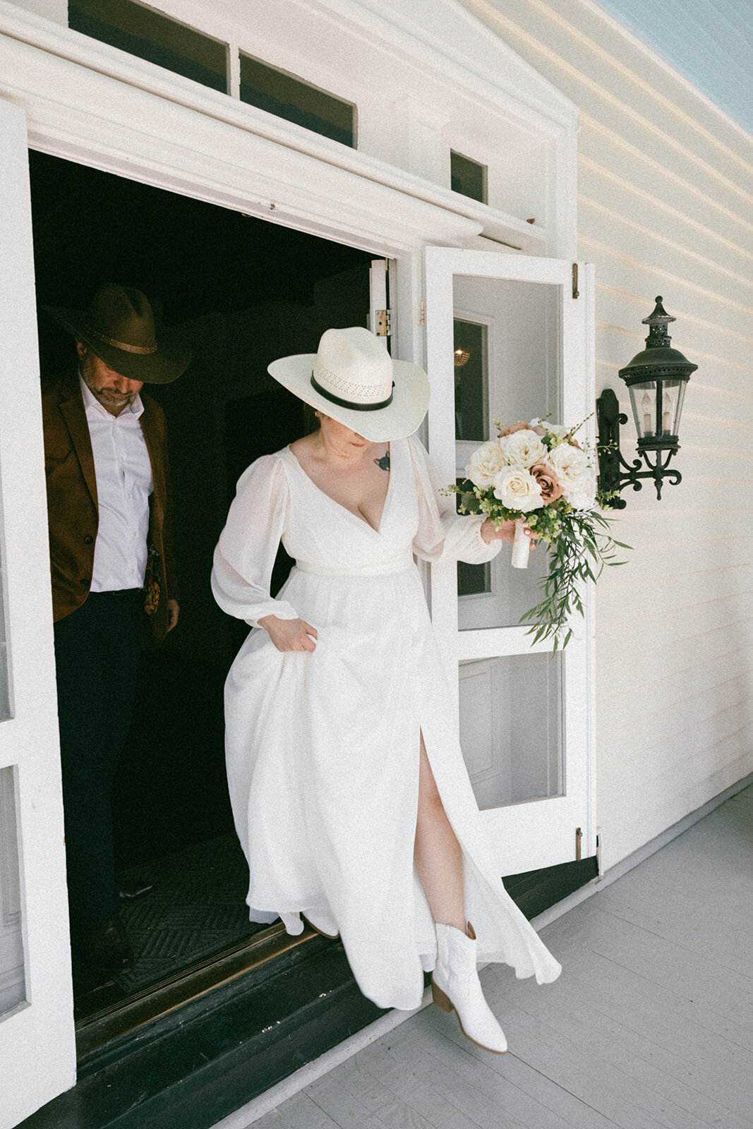 Bride candidly steps out of the house in her white cowboy hat and white dress onto a sunlit porch. 