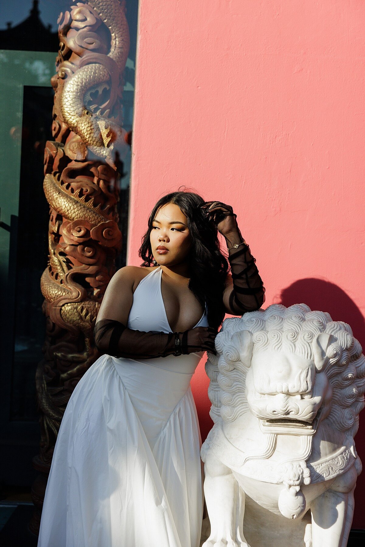 Bold editorial senior portrait of a teen girl in a white halter dress in Chinatown Los Angeles, leaning on a stone lion statue with vibrant architecture