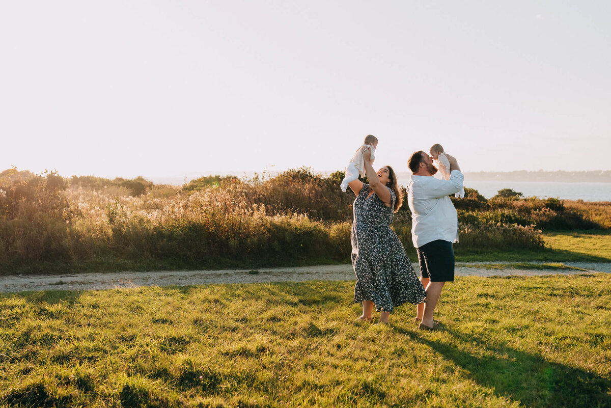 A mother and father stand back to back and hold their twin daughters up in the air at sunset during their photoshoot with Allison Wolf, Boston family photographer