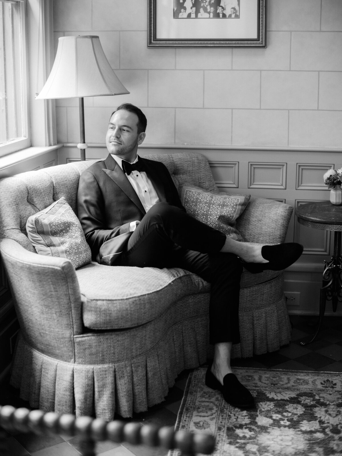 The groom sitting relaxed on a cozy couch dressed in formal attire, embodying a mix of calm elegance and preparation before the exciting moments of his wedding day unfold.