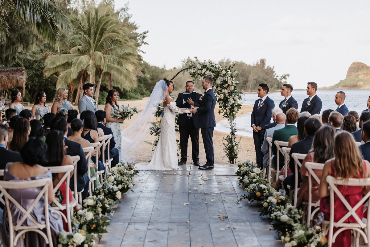 Bride and groom ceremony at Kualoa Ranch on the beach, holding hands with palm trees and ocean in the background