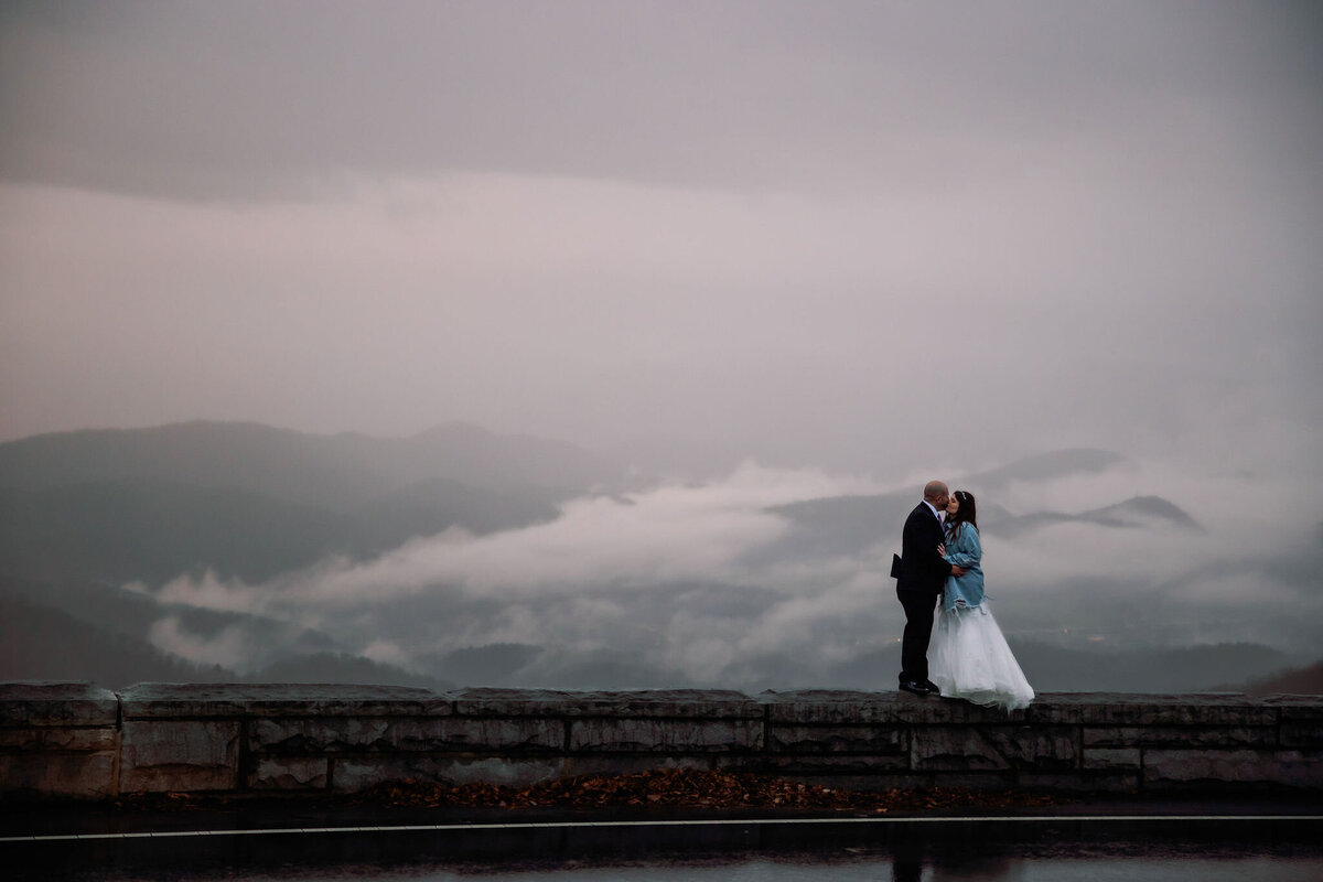 cloudy day in the Smoky Mountains with bride adn groom embracing at foothills parkway overlook with clouds hanging over the mountains