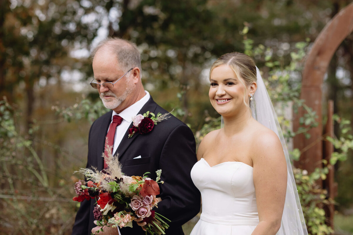 father of the bride walking his daughter down the aisle at her outdoor wedding venue in Charlottesville with a lush wood surrounding them while the bride smiles at her groom