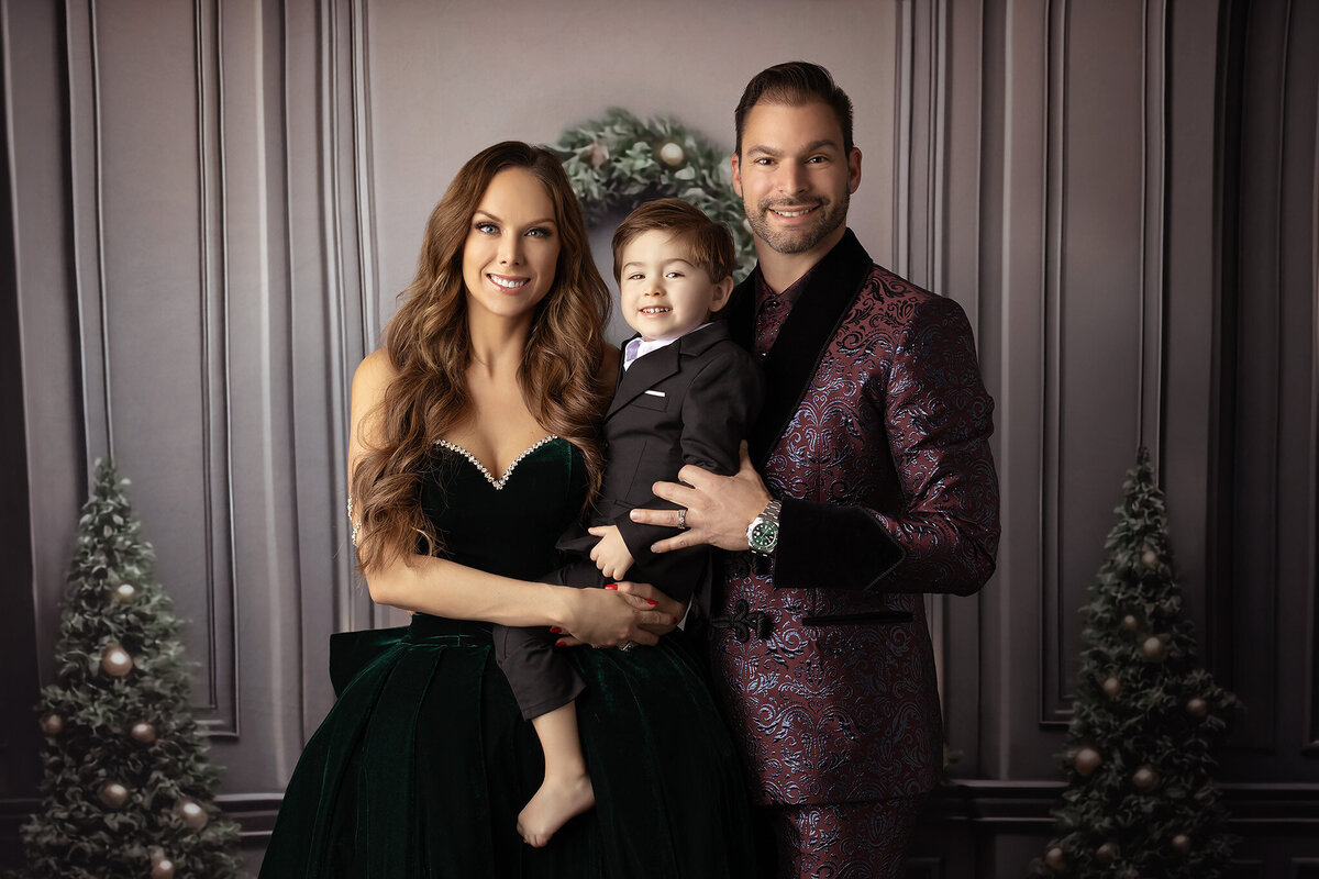 Elegant family portrait with a mother in a green velvet gown, father in a patterned suit, and their son in a black tuxedo, captured by a Dallas family photographer during a holiday session.