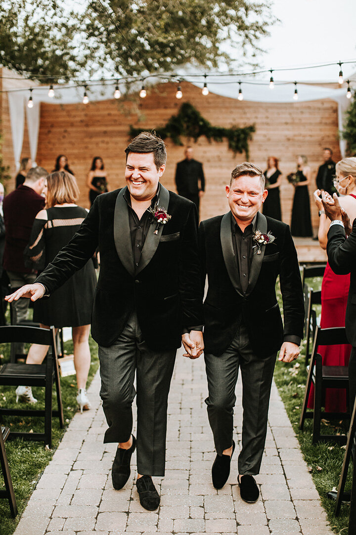 Two grooms wearing black tuxedos walk down the aisle at their wedding, smiling and holding hands.