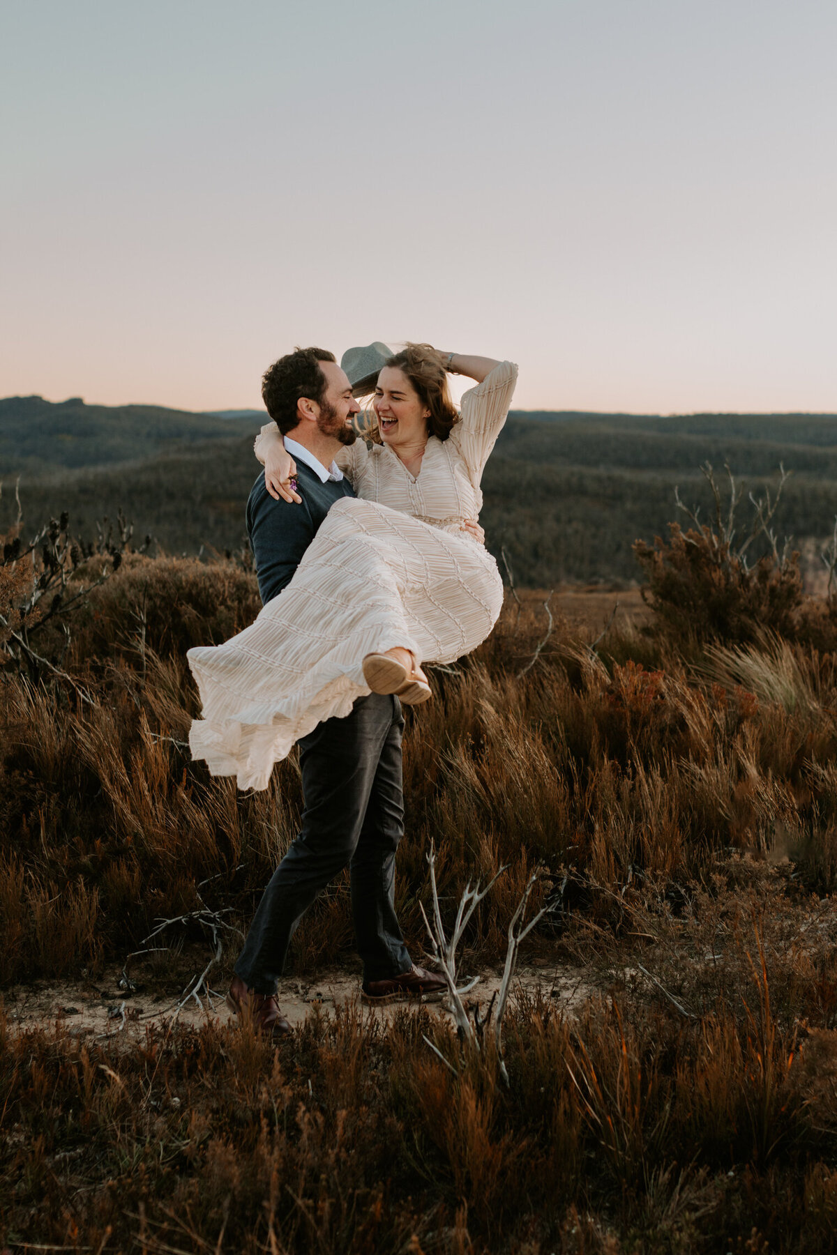 Father lifting and carrying mother while both laugh during a playful family session in the Blue Mountains at sunset, surrounded by native Australian bushland.