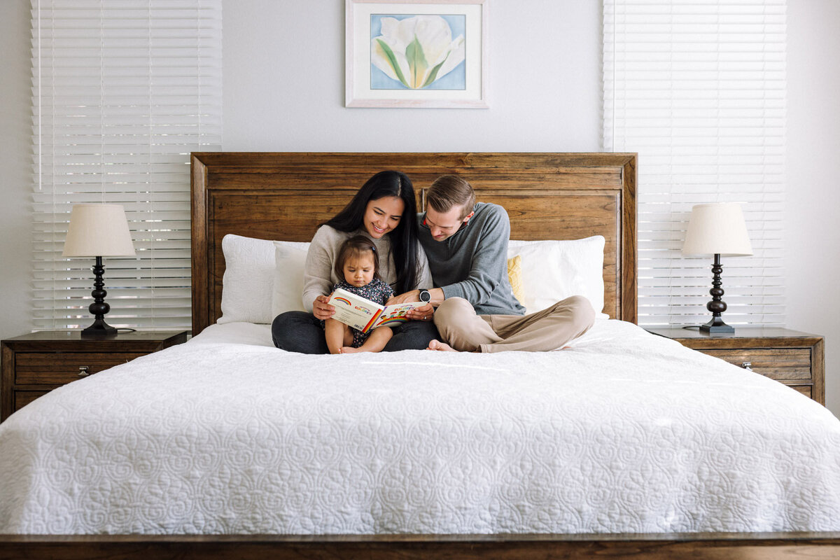 parents and toddler daughter sitting on white bed reading together