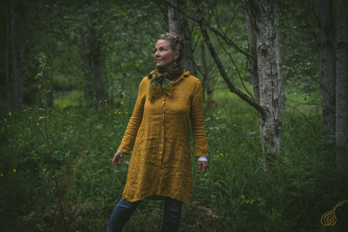 A woman wearing a bright yellow dress poses in a forest, surrounded by trees and the peaceful ambiance of the outdoors.