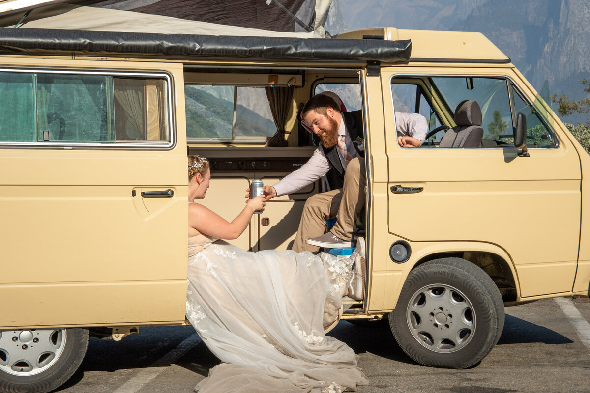 A groom reaches for a can of beer in a yellow van as his bride sits on the sliding door step of the van on their Yosemite elopement day.