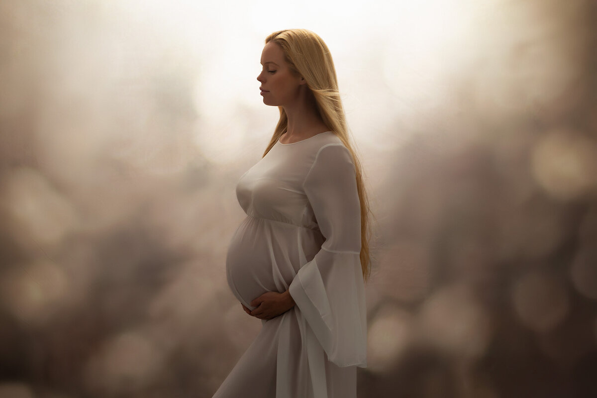 A serene woman in a flowing white gown stands gracefully against a softly blurred golden background, her long blonde hair cascading gently over her shoulder. The soft, glowing light highlights the beauty of DFW maternity photography by Lindsay Walden.
