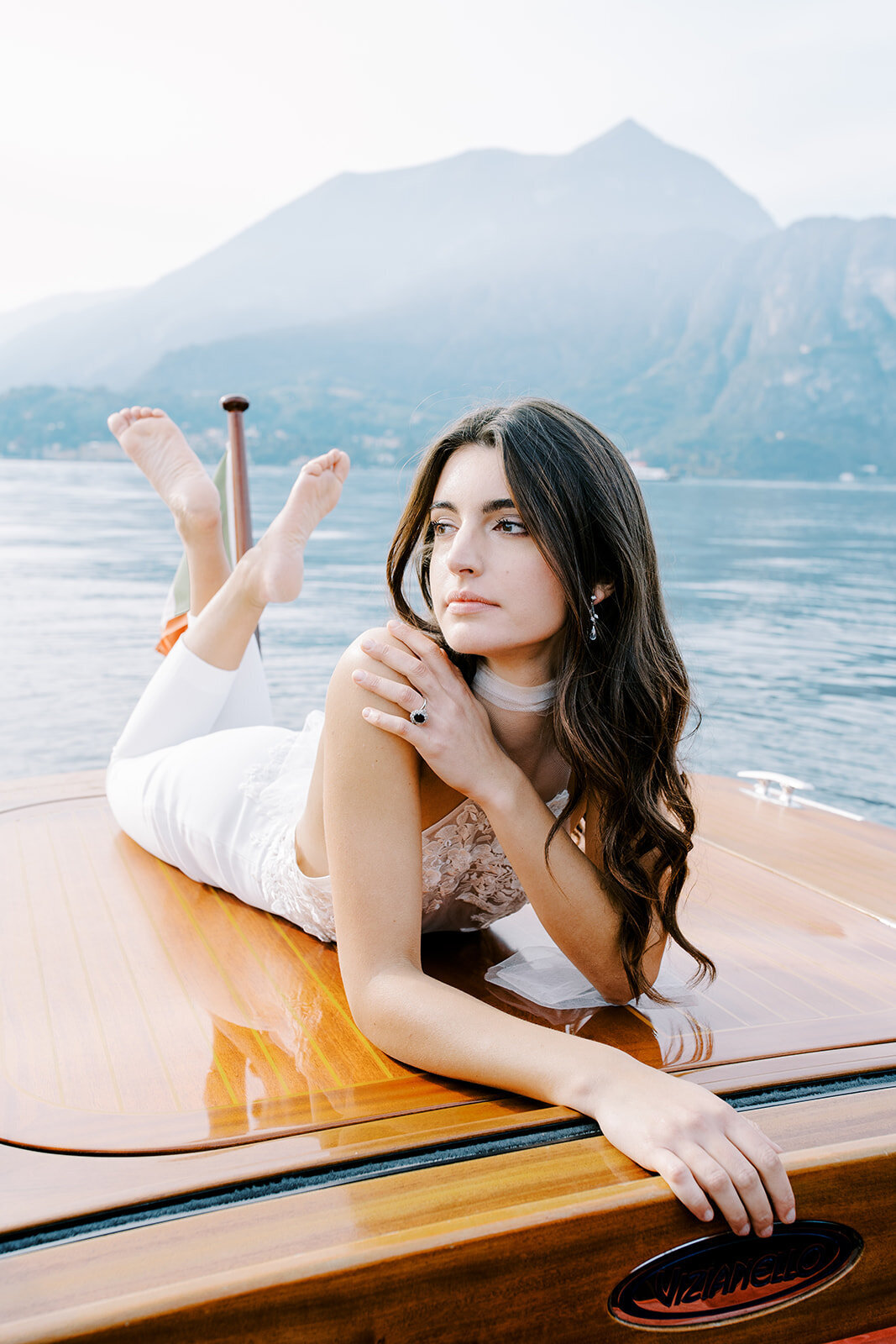 Film photograph of bride and groom on a wooden boat on Lake Como in Italy she is wearing a white halter jumpsuit with her hair down in loose waves and he is wearing grey and navy cashmere sweater and coat photographed by Italy wedding photographer