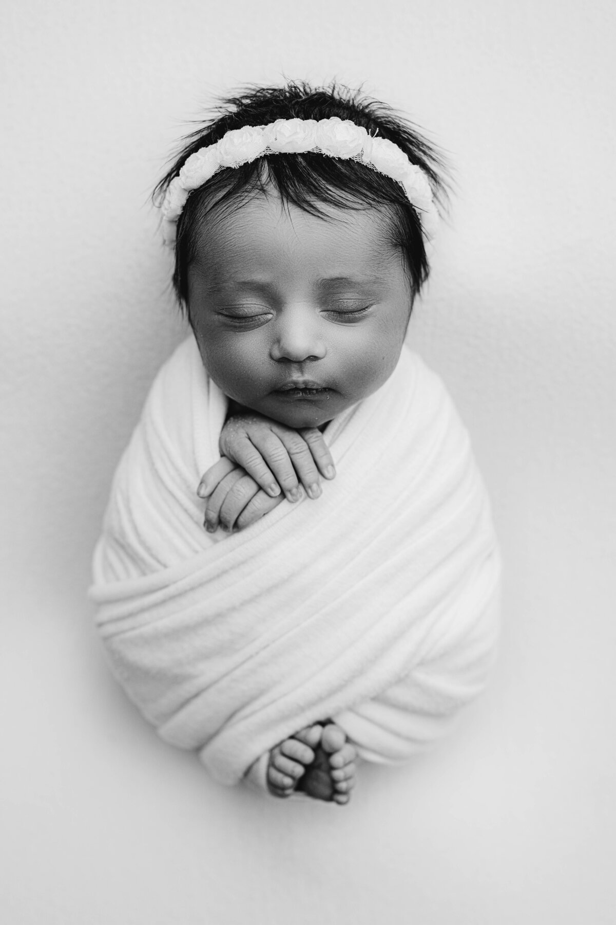 Black and white image of a swaddled newborn girl with floral headband, hands crossed over chest.