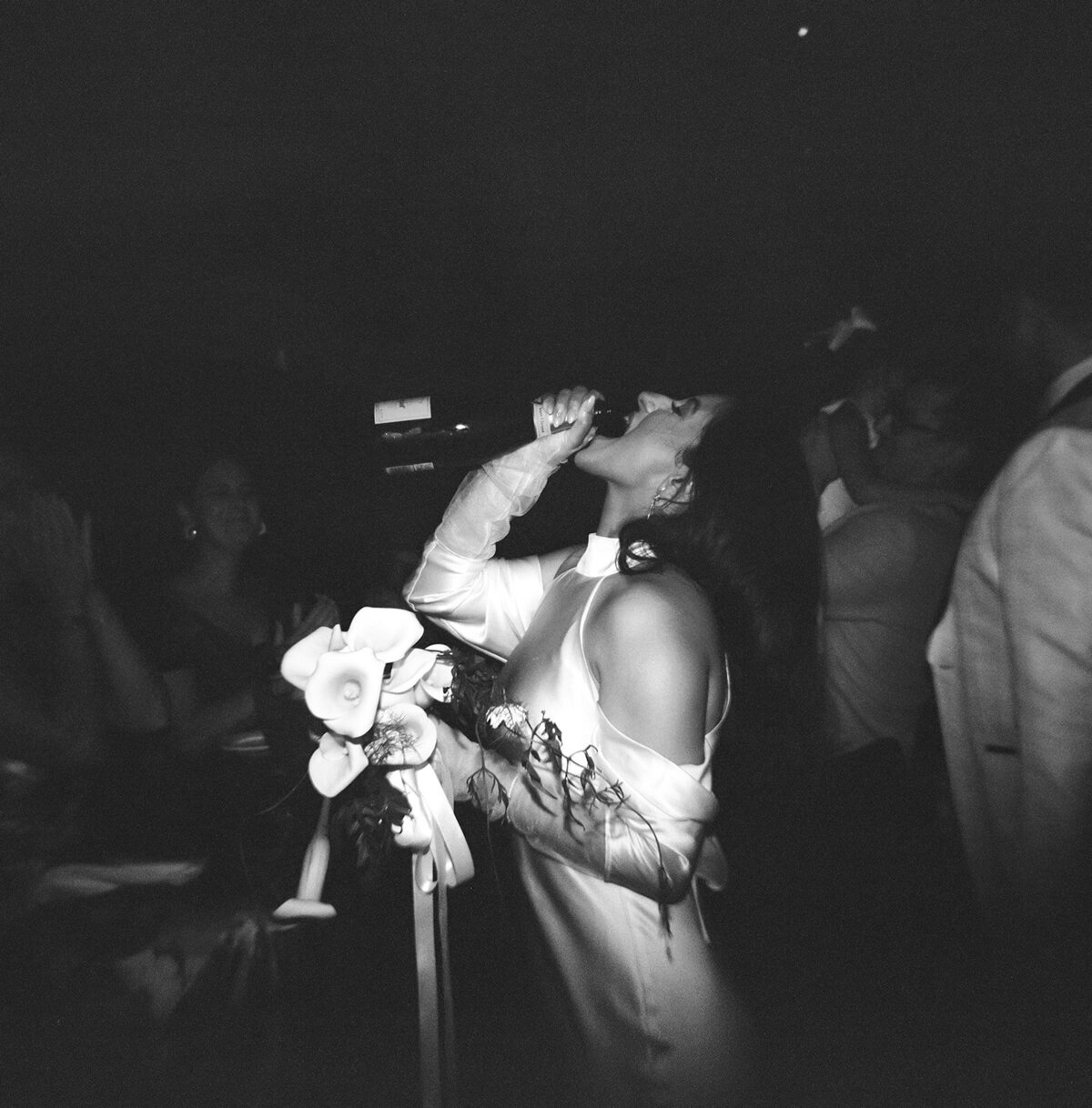 Black and white film photo of a bride singing into a microphone while holding her bouquet during a lively New York City elopement reception.