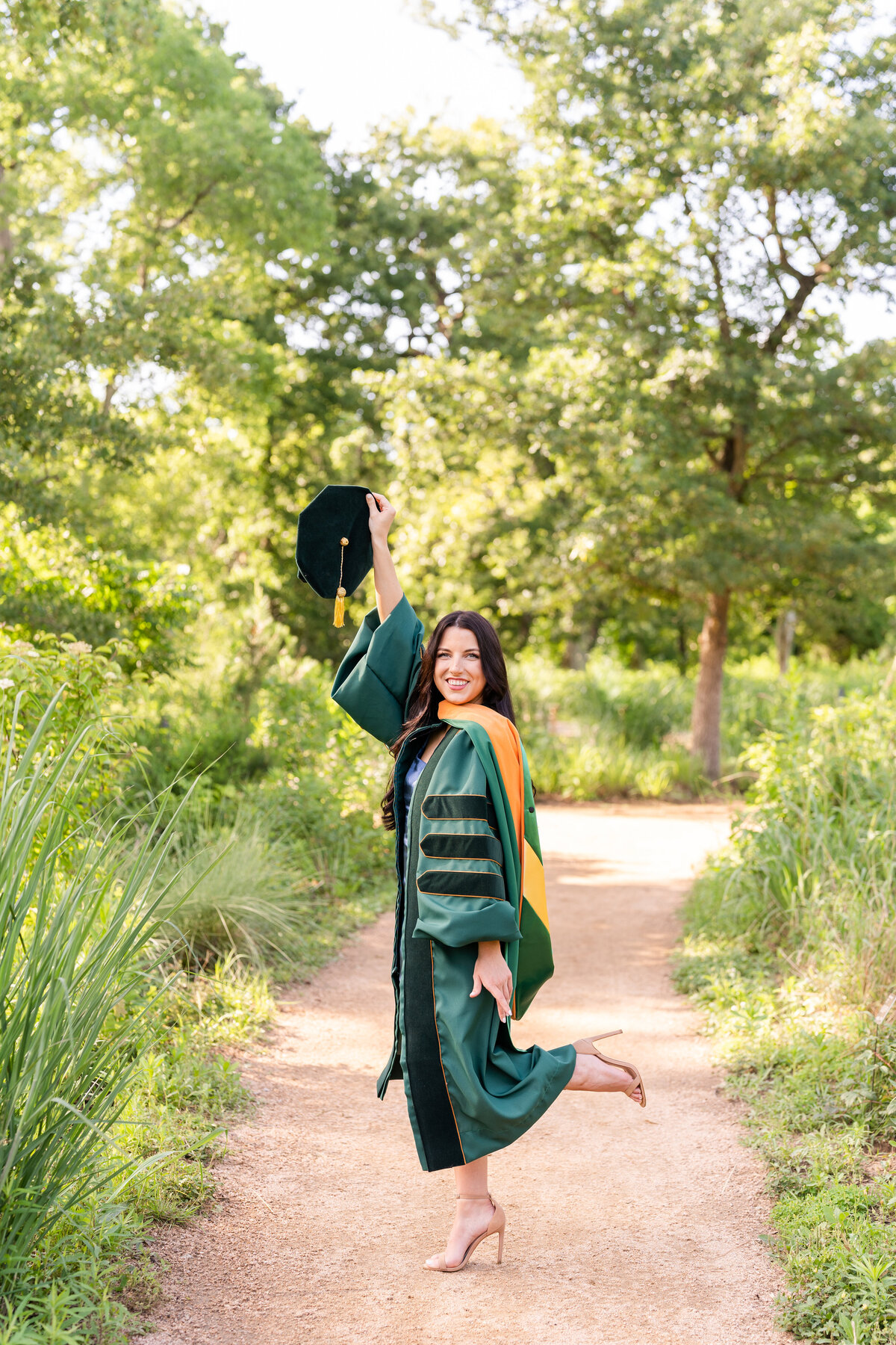 Baylor senior girl wearing doctorate gown, hood and holding up hat while celebrating on pathway surrounded by nature of Houston Arboretum