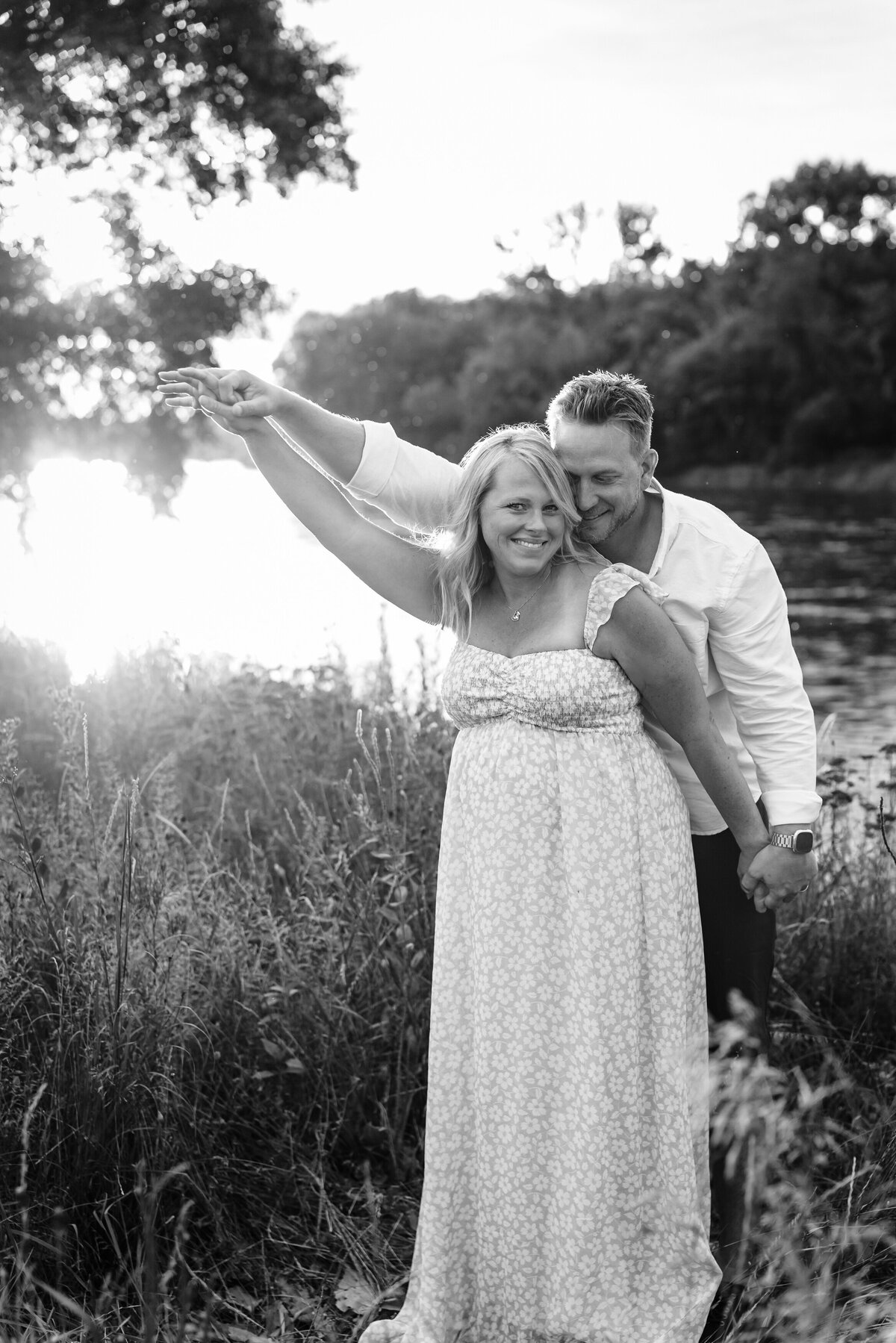 A black and white photo of the couple near a river, with the man gently holding the woman from behind.