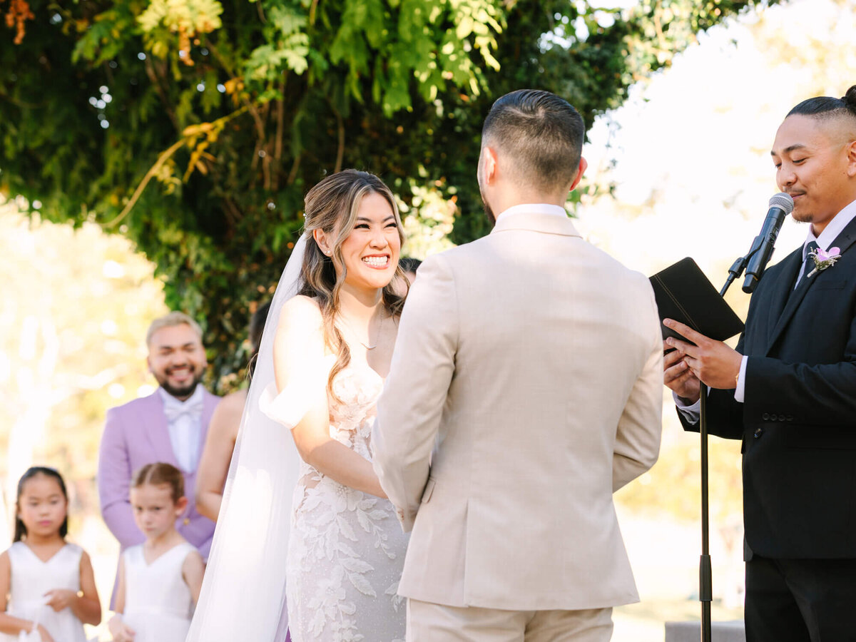 Bride in a white gown smiles warmly at groom during outdoor wedding ceremony. Officiant reads from a book. Bridesmaids in white and a groomsman in lavender watch under a tree.