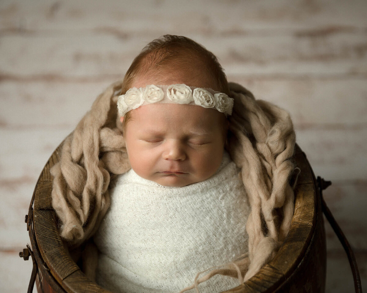 Sleeping newborn girl wrapped in a white wrap with a lace headband. She is in a wooden bucket with brown colored wool.