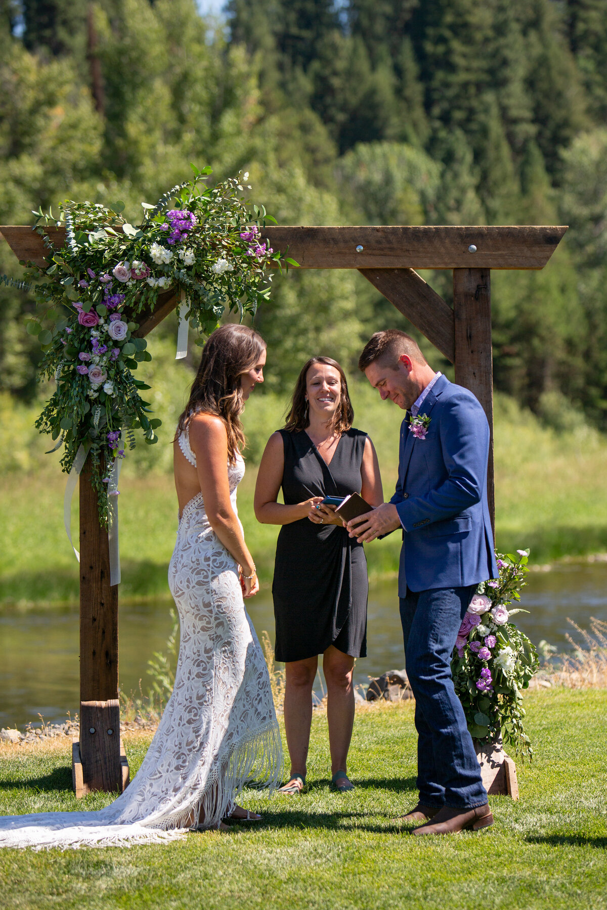 A bride and groom stand facing each other while their officiant stands behind them under a wooden arch with flowers.