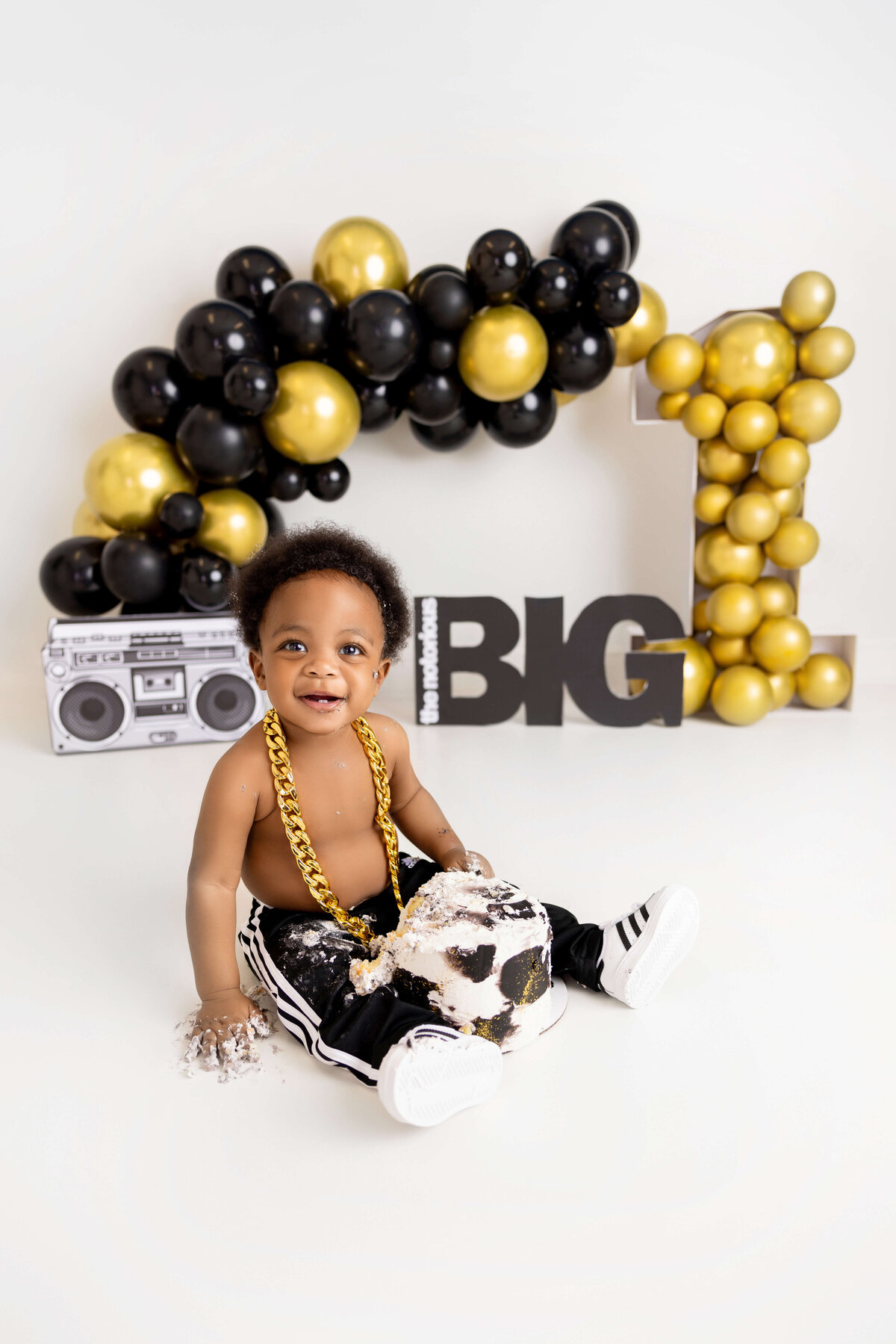 A baby in black and white pants with a gold chain sits amidst a smeared cake. Behind, a black and gold balloon arch forms the number "1", accompanied by a toy boombox and the word "BIG". Captured perfectly by Jacksonville Cake Smash Photography, it’s a delightful scene of first birthday joy.