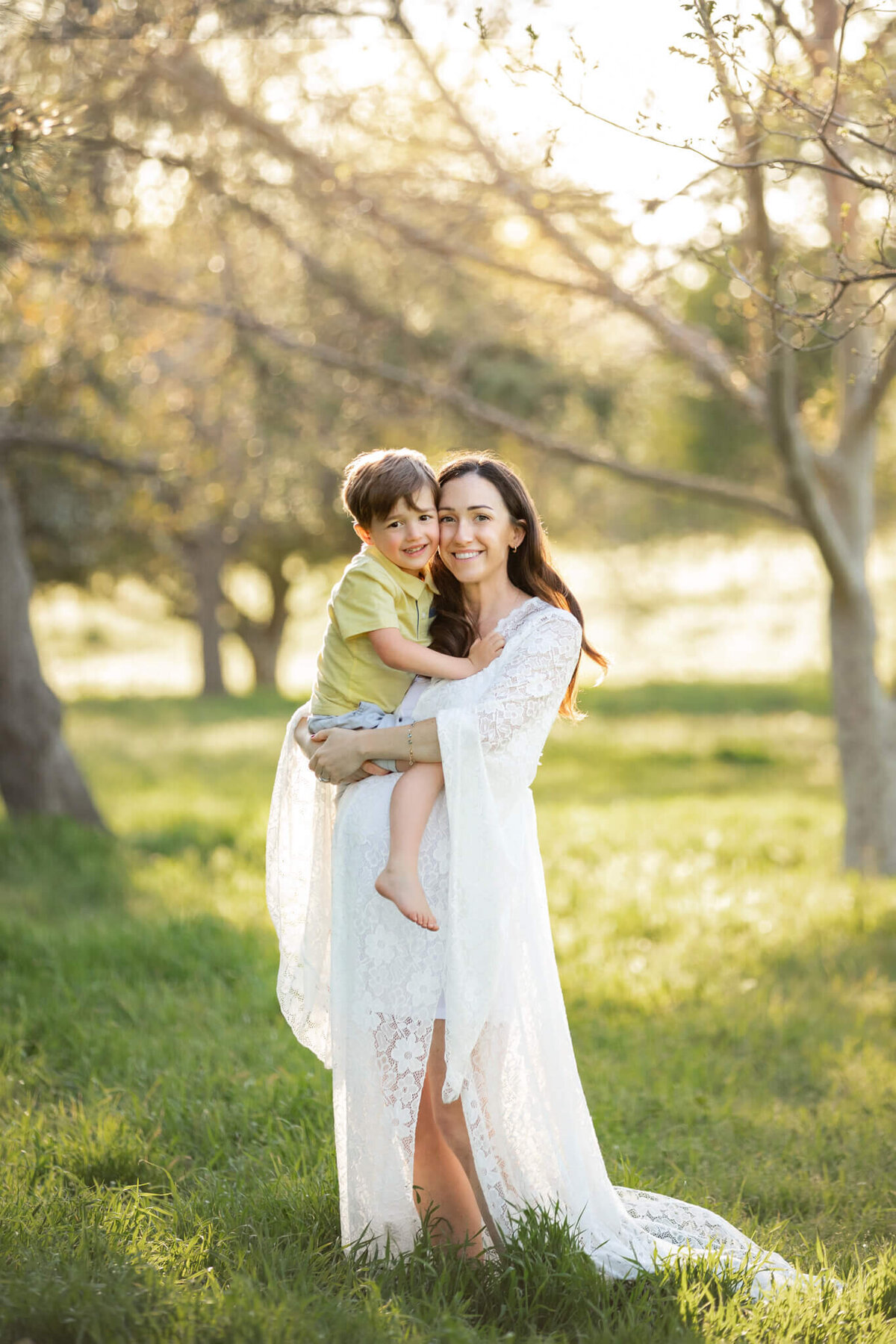 Pregnant mom wearing a white dress in the park with her first son wearing a yellow polo shirt and hugging