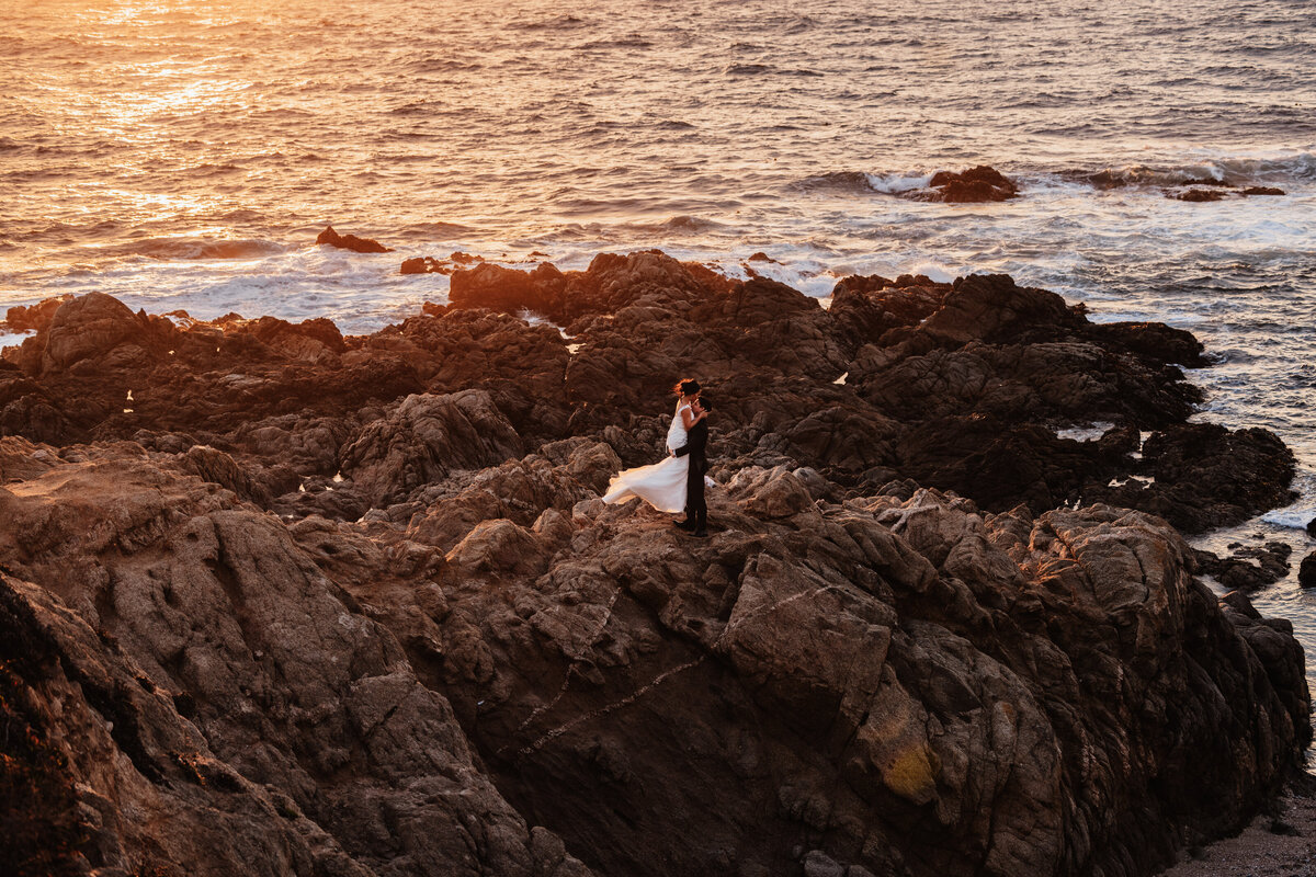 Shannon Lee, from Love Like Wild Photography, captures a couple embracing on rocky cliffs at sunset by the ocean.