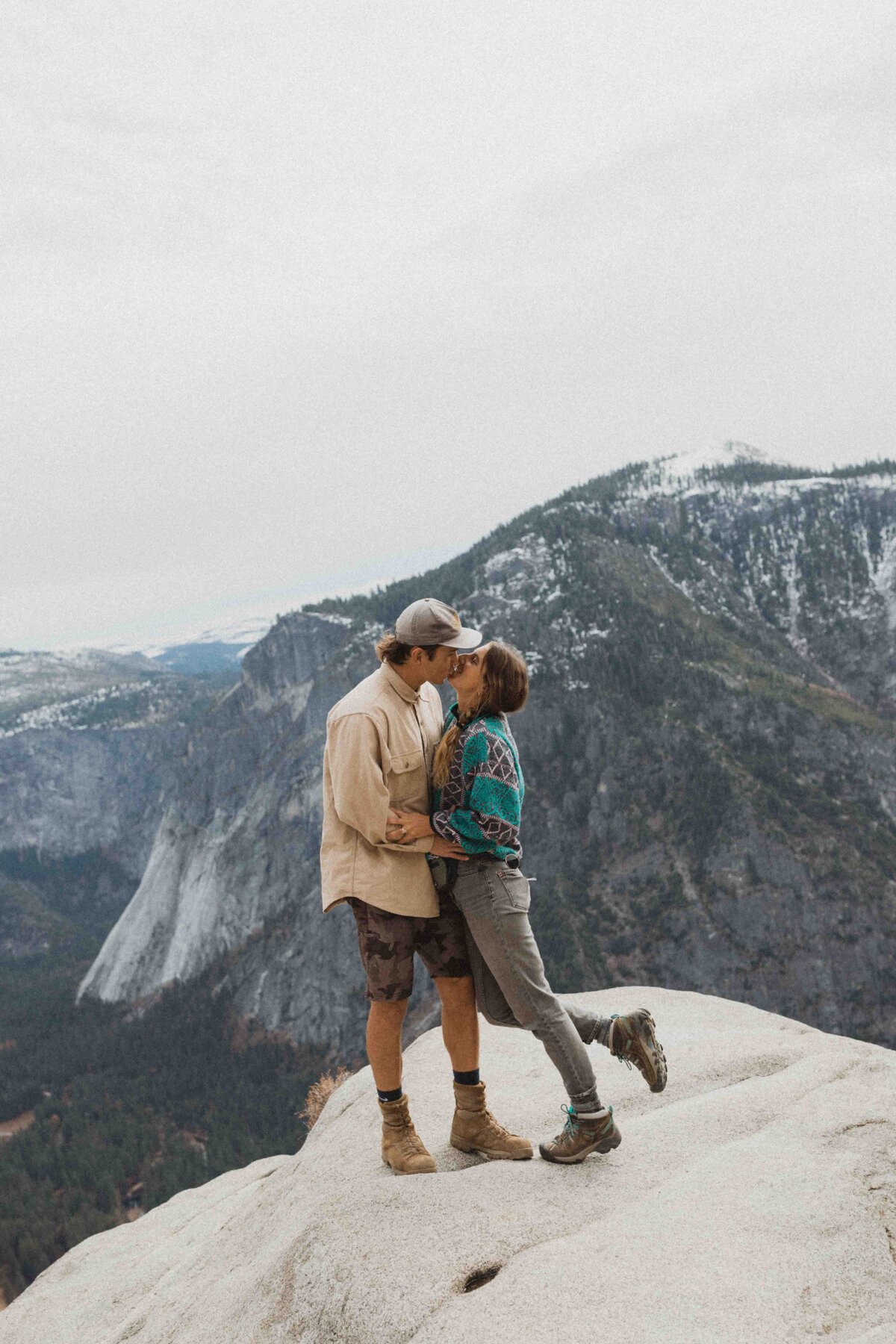 Couple standing and kissing on top of mountain in Yosemite National Park. Girl leaning back, with foot popped. Both dressed in hiking gear.