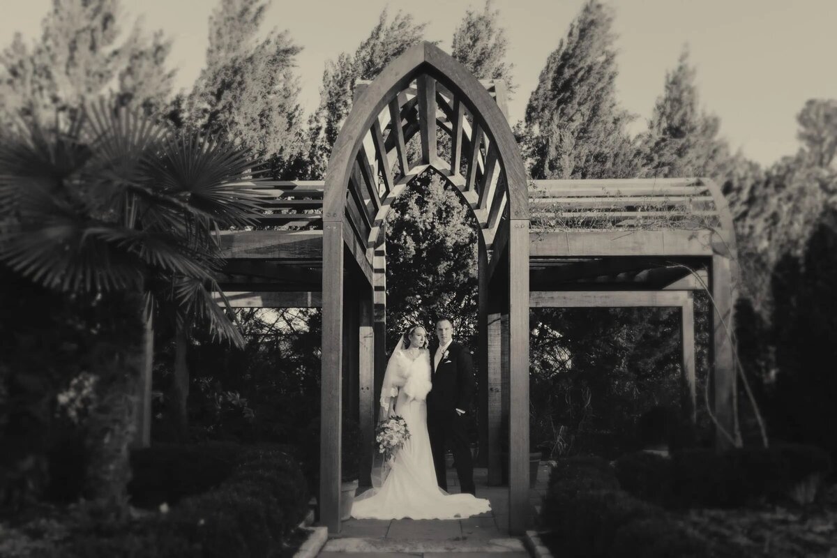 A couple stands in a vintage black and white photo under an ornate garden arch
